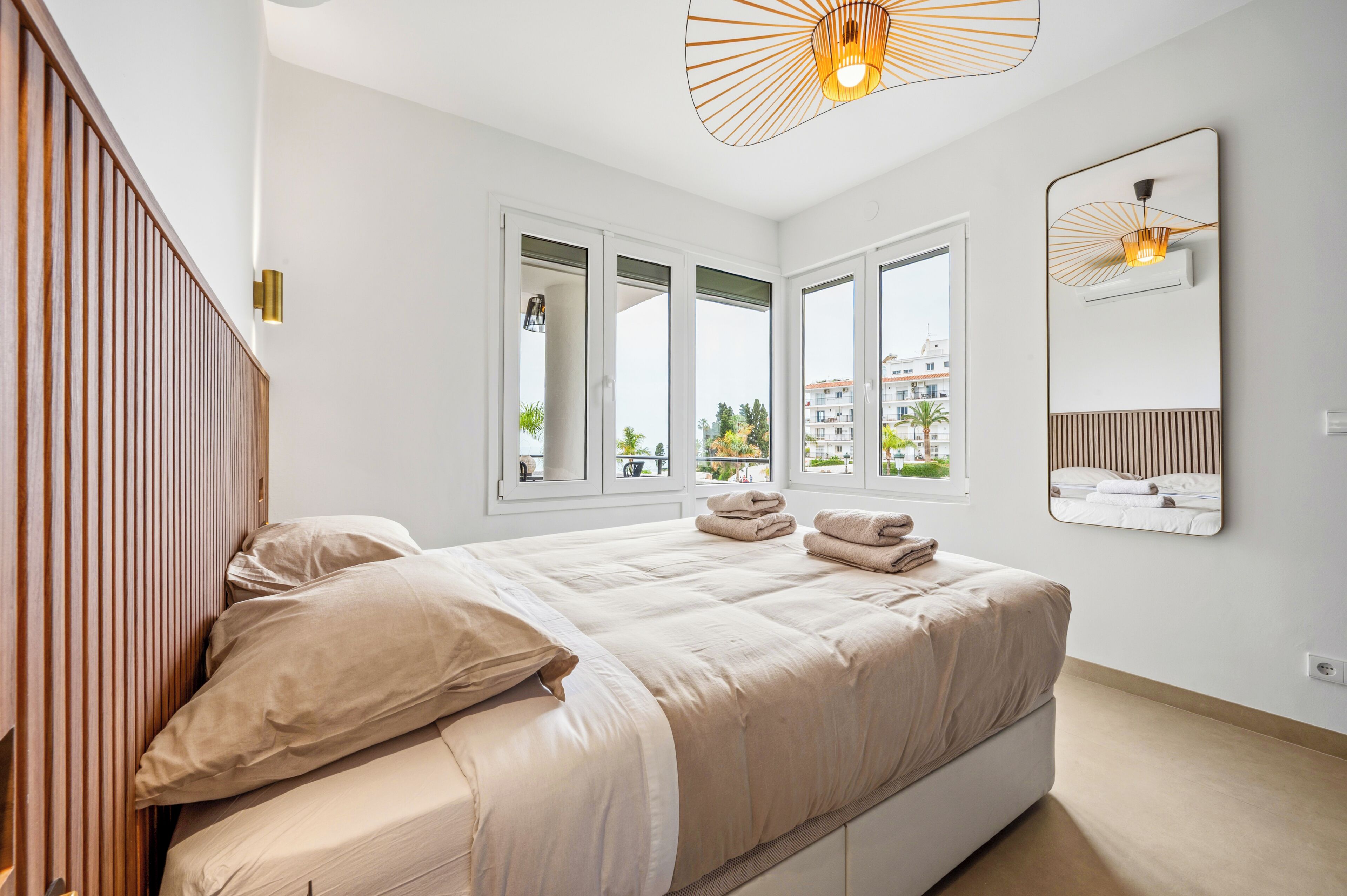A modern bedroom with a large bed, multiple windows, a wooden headboard, and a wall-mounted mirror.