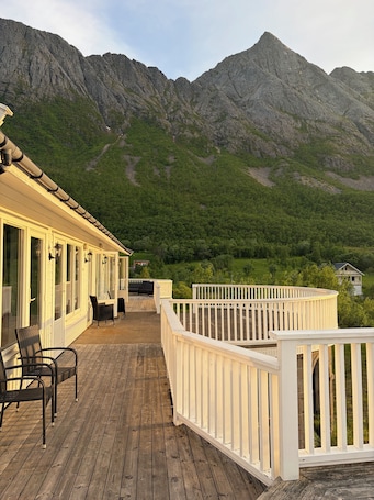 A wooden deck with outdoor furniture, a white railing, and a mountain backdrop.. Senja Living Sea View apartment