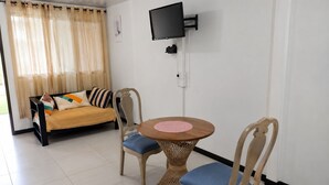 Une pièce comprenant un canapé, deux fauteuils, une table ronde, un téléviseur fixé au mur et une fenêtre avec des rideaux.