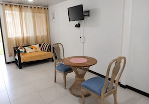 Ein Zimmer mit einer Couch, zwei Sesseln, einem runden Tisch, einem an der Wand befestigten Fernseher und einem mit Vorhängen drapierten Fenster.