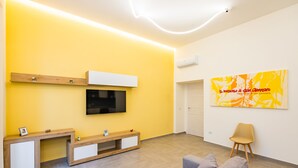 A modern living room with a yellow wall, a mounted TV, a wooden console table, a grey sofa, and a large abstract painting. - Solaris Studio - Solaris Suites (Naples)