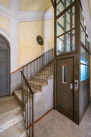 Una escalera con barandales metálicos, una puerta con una luz de emergencia roja y un muro con textura.