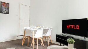 Een kamer met een eettafel, stoelen, een televisie waarop 'NETFLIX' wordt weergegeven, en een kamerplant.