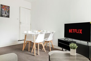A room with a dining table, chairs, a TV displaying 'NETFLIX,' and a potted plant. - NEW! 4BR House, free parking, near City Centre (Nottingham)