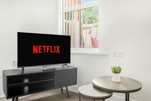 A modern living room with a flat-screen TV displaying 'NETFLIX,' a black entertainment unit, a small round table with a potted plant, and a window with a view of a brick wall and greenery. - NEW! 4BR House, free parking, near City Centre (Nottingham)
