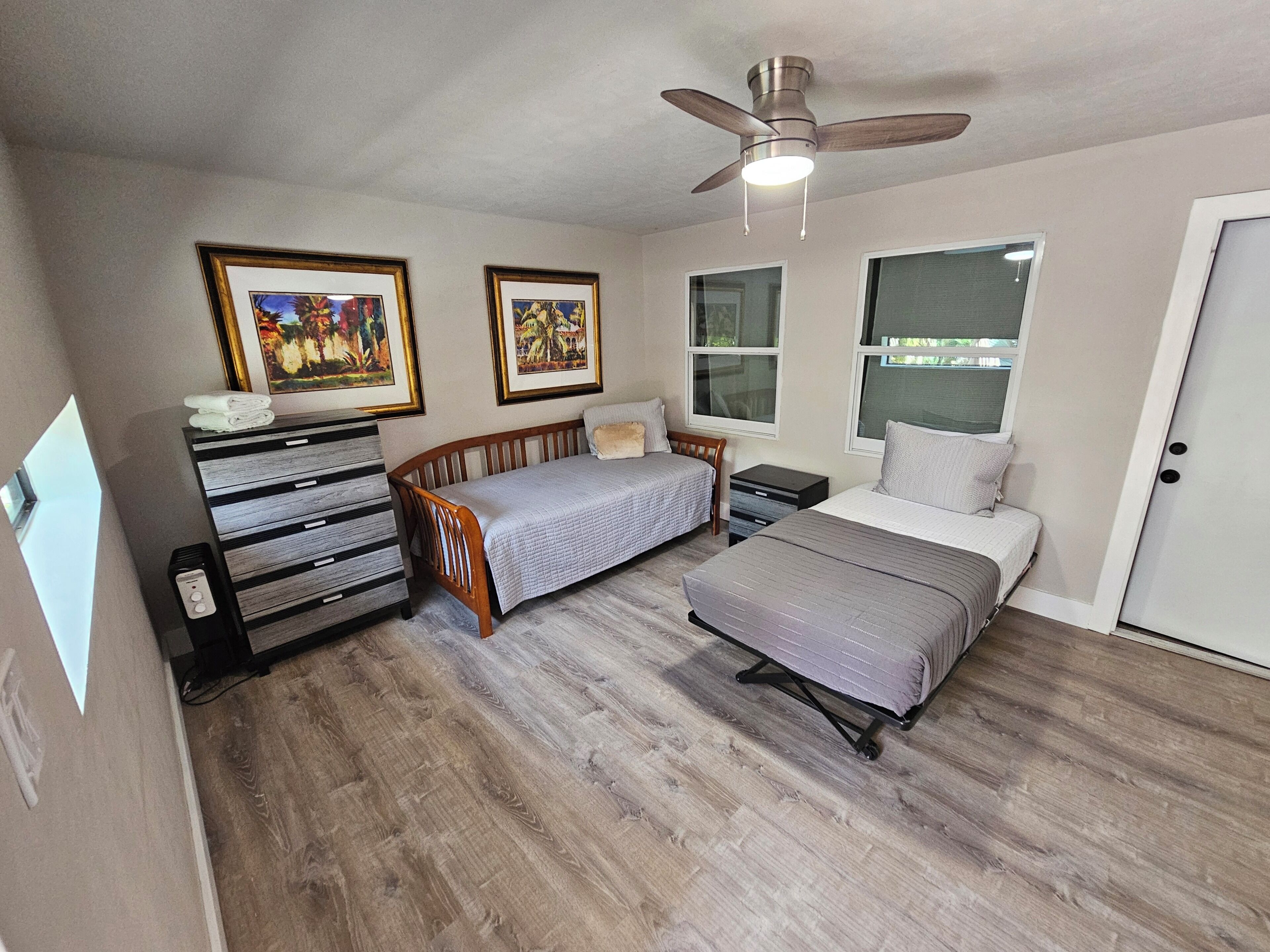 A bedroom with two beds, a ceiling fan, framed artwork, a dresser, and a window.
