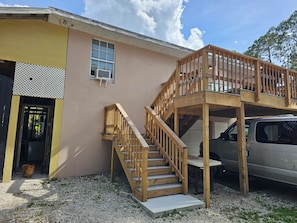 A two-story building with a wooden staircase and balcony, a parked car, and a door leading to a covered entrance. - Guest house pine ridge 2nd floor unit over shop. (Naples)