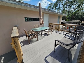 A deck with a table and chairs, a wooden railing, and a building with a window and door. - Guest house pine ridge 2nd floor unit over shop. (Naples)