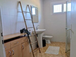 Une salle de bain avec des carreaux blancs, un meuble en bois avec des tiroirs, un lavabo, un bidet, une échelle et une fenêtre.
