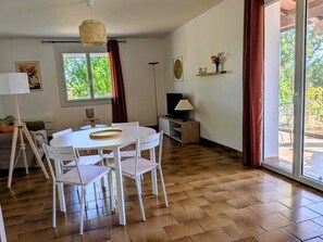 Un coin repas avec une table et des chaises blanches, une lampe, un téléviseur et une fenêtre avec des rideaux.