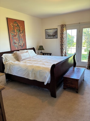 A bedroom with a bed, a nightstand, a lamp, a wooden chest, a painting on the wall, and a window with curtains.