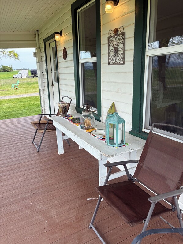 A covered porch with a bench, chairs, and a table with various items.