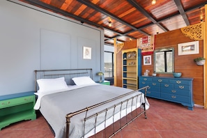 Quarto com estrutura de cama em metal, mesa de cabeceira verde, cómoda azul e parede de madeira com detalhes decorativos.