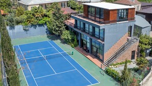 A tennis court surrounded by a modern building with a glass facade, a staircase, and a small garden area.