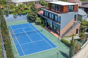 A tennis court surrounded by a modern building with a glass facade, a staircase, and a small garden area.