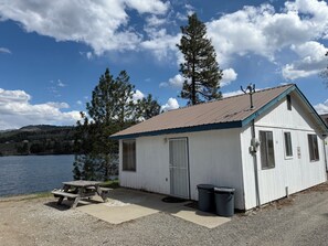 Une petite cabane blanche avec un toit en métal, une table de pique-nique et deux poubelles.