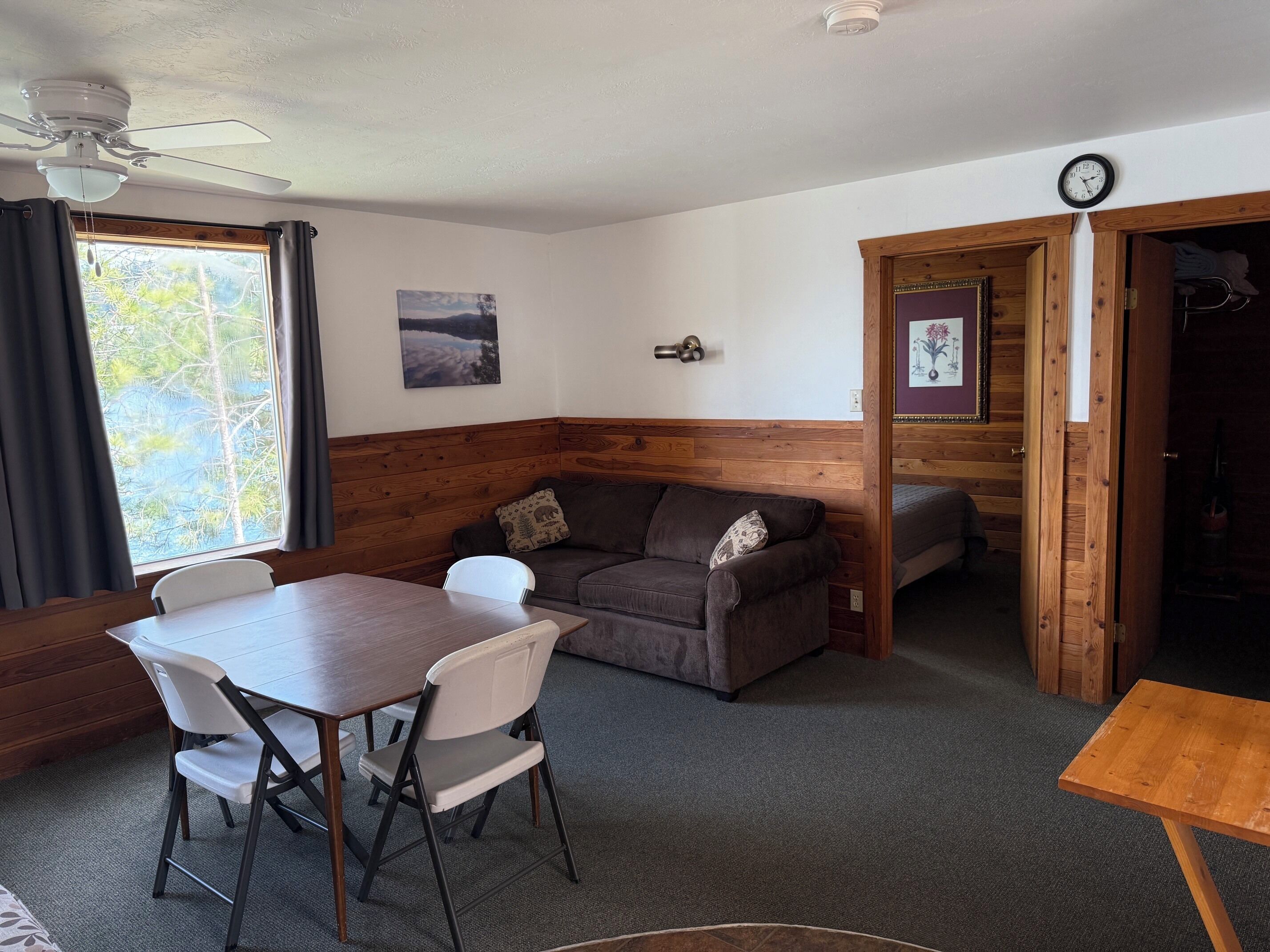 A room with a wooden bench, a table, and a sofa. There is a ceiling fan, a clock, and a window with curtains.