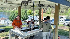 A covered outdoor area with a table, people working on a project, and a lake in the background.