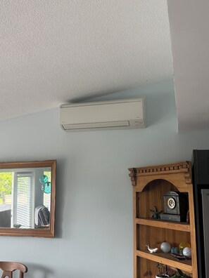 Et værelse med en loftmonteret aircondition, en træbogreol med pyngegenstande og et spejl der reflekterer et vindue med persienner.