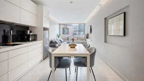 Una cocina moderna con armarios blancos, un comedor con una mesa blanca y sillas grises, y un ventanal con vistas a la ciudad.