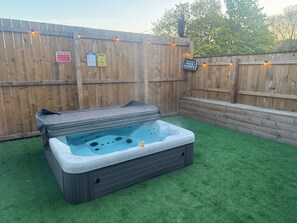Ein Whirlpool in einem Hinterhof mit Holzlattenzaun, einem Schild mit der Aufschrift 'Willkommen in unserem Biergarten' und einem Warnhinweis zu rutschigen Oberflächen.