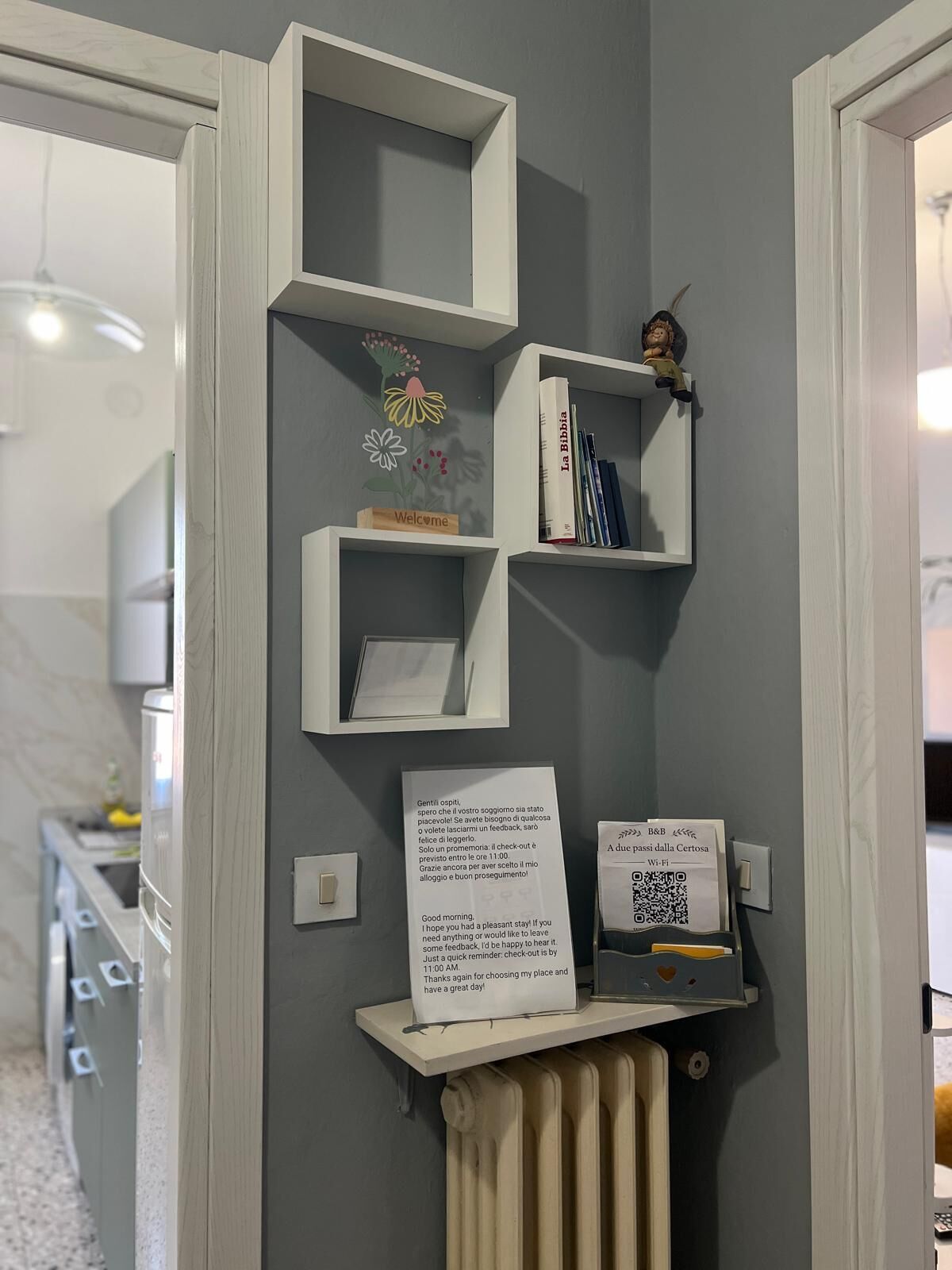 Una habitación con pared gris, una puerta blanca y una estantería de marco blanco con un letrero de bienvenida, una pequeña figurita y un código QR.