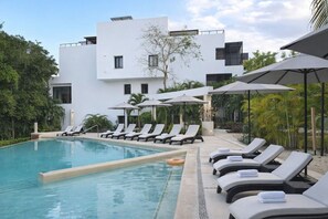 Ein Schwimmbecken mit Liegestühlen und Sonnenschirmen, umgeben von einem modernen Gebäude mit flachem Dach und wenigen Fenstern.