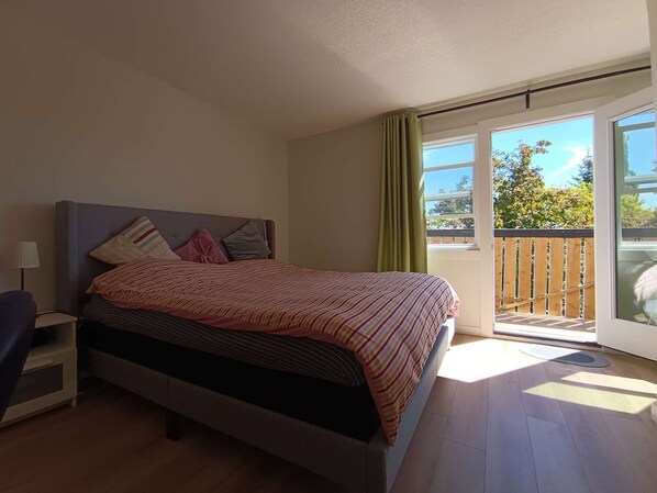 Un dormitorio con cama, mesita de noche, una silla, ventana y balcón.