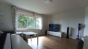 A living room with a piano, a TV, a vacuum cleaner, and a window with curtains.