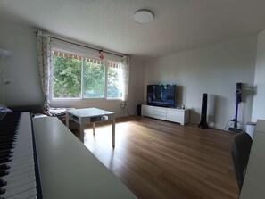 A living room with a piano, a TV, a vacuum cleaner, and a window with curtains.