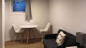 A room with a round table, two chairs, a sofa, a small bedside table, and a window with blinds.