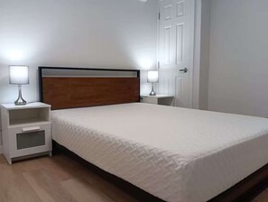 Una cama bien tendida con un cabecero de madera, dos mesitas de noche con lámparas y una puerta blanca.