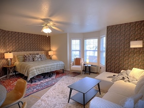 A bedroom with a bed, two bedside tables, a chair, a small table, a ceiling fan, and a window with blinds.