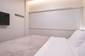 Habitación de hotel minimalista con cama, televisor de pantalla plana en la pared y ventana con persiana blanca.