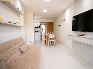 A modern living room with a sofa, a dining area with a table and chairs, a kitchen with appliances, and a flat-screen TV mounted on the wall. - Cond em Resort Pe na Areia - By MME (Maceió)
