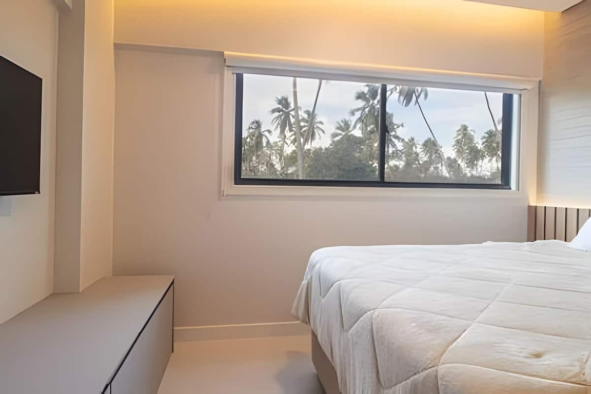 Un dormitorio con cama, ventana con vista a palmeras, televisor de pantalla plana en la pared y una mesita de noche.