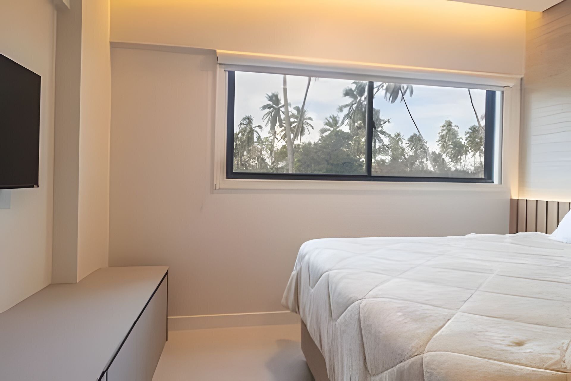 A bedroom with a bed, a window with a view of palm trees, a flat-screen TV on the wall, and a bedside table.