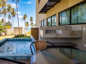 A modern outdoor pool area with a clear view of the ocean and palm trees, a glass table, and a built-in stone shelf. - Cond em Resort Pe na Areia - By MME (Maceió)