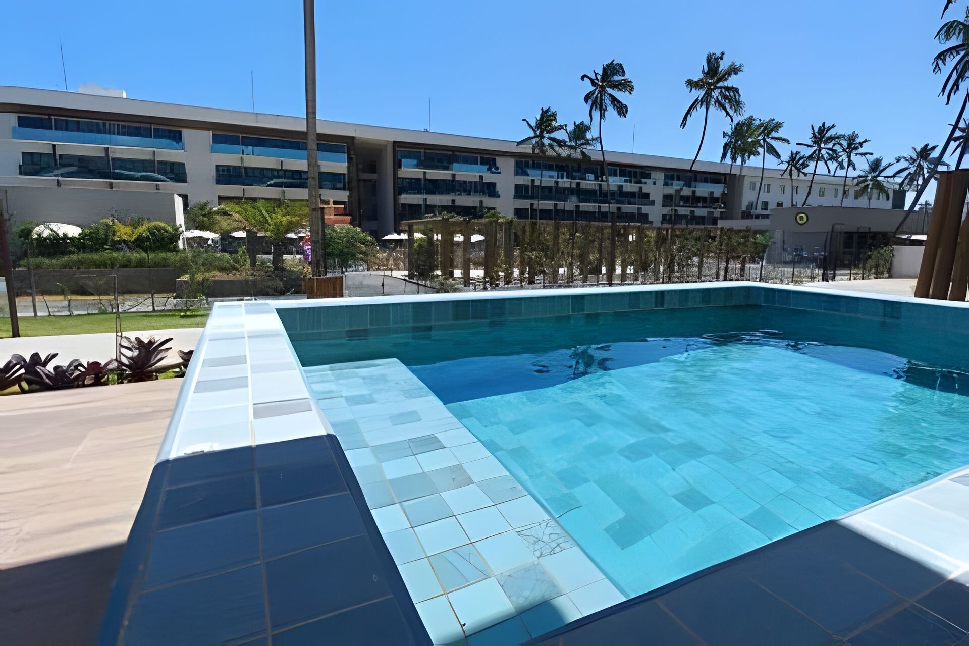 A swimming pool with a tiled edge, surrounded by a building with multiple floors and palm trees.
