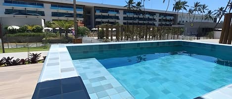 A swimming pool with a tiled edge, surrounded by a building with multiple floors and palm trees.