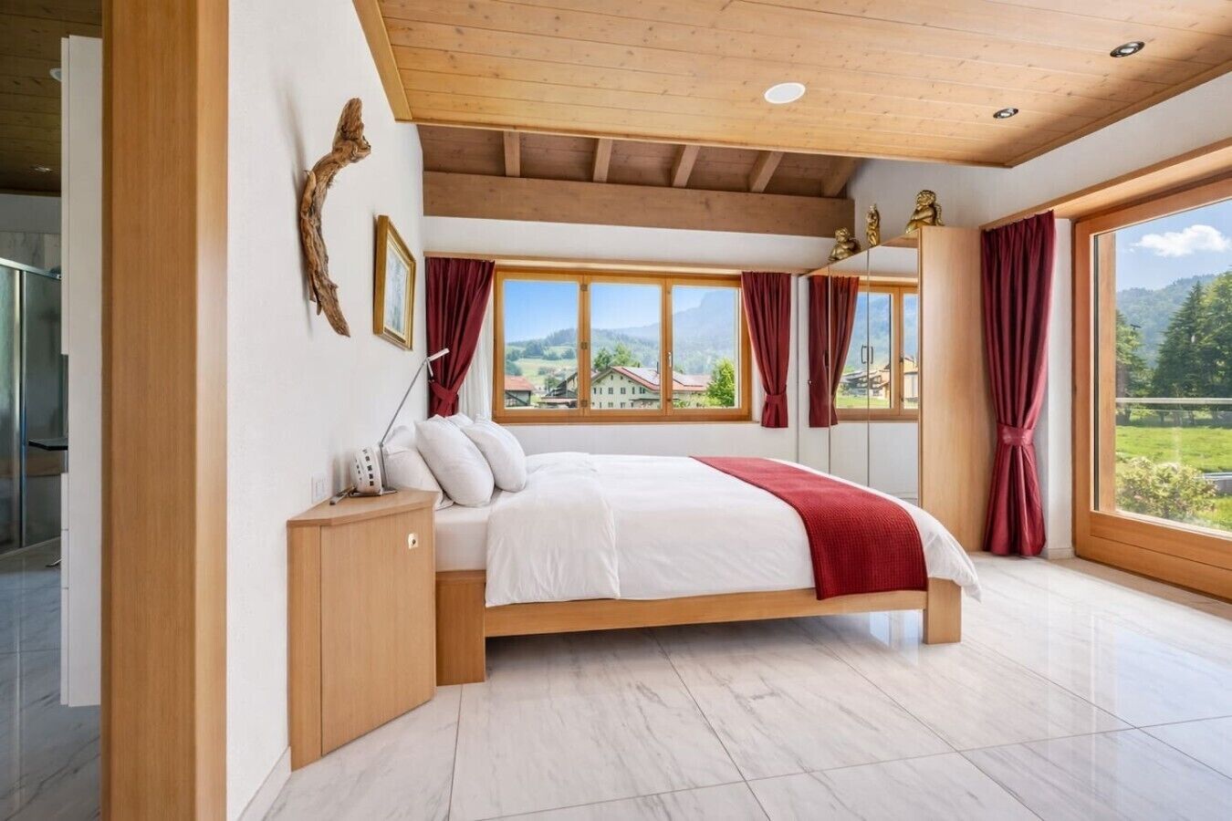 Een slaapkamer met een bed, houten hoofdeinde, een nachtkastje, een lamp en uitzicht op een groen landschap door grote ramen.