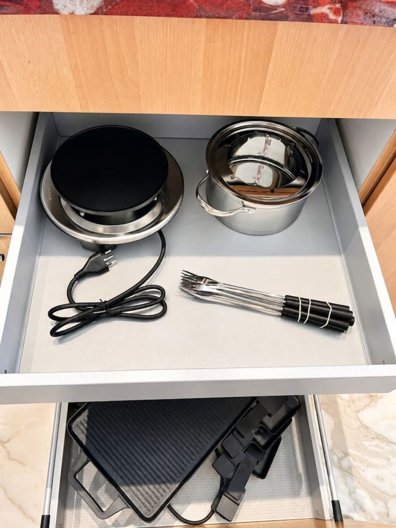 A drawer with a black electric kettle, a stainless steel pot, a black cord, and a set of chopsticks.