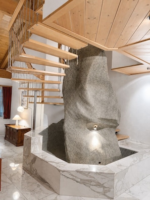 A modern interior with a wooden staircase, a large stone sculpture, and a marble floor.