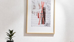 Ein gerahmtes Foto einer verschneiten Landschaft mit Ski an einer Wand, eine Topfpflanze und ein Holzkabinet mit Stühlen.