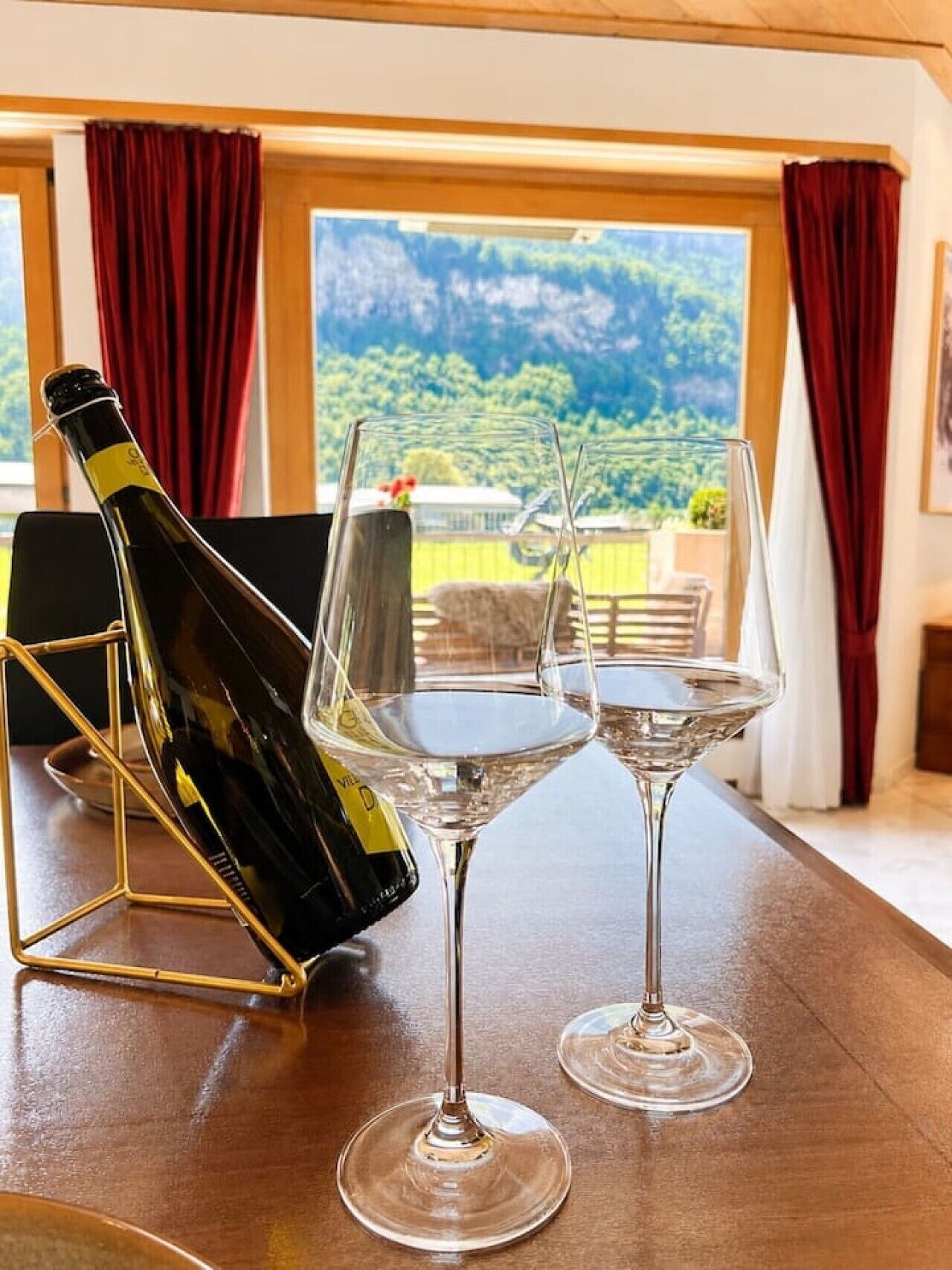 A table with a bottle of wine and two glasses, set against a backdrop of a scenic view through large windows.