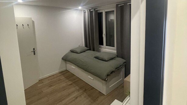Um quarto com uma cama, mesa de cabeceira, janela com cortinas e uma porta.