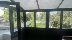 Quarto com piso de madeira, um banco e grandes janelas com vista para um jardim e edifícios.