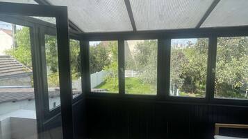 Une pièce dotée d’un plancher en bois, d’un banc et de grandes fenêtres donnant sur un jardin et des bâtiments.