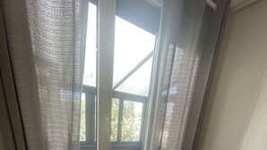 Quarto com escrivaninha de madeira, cadeira, radiador, janela com cortinas e lixeira.