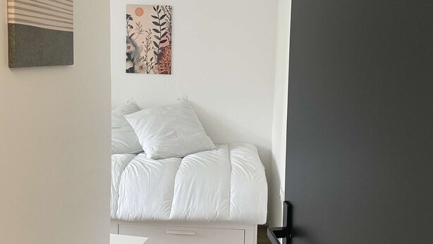 Quarto com uma cama, uma mesa de cabeceira branca, uma parede com duas obras de arte e uma porta.
